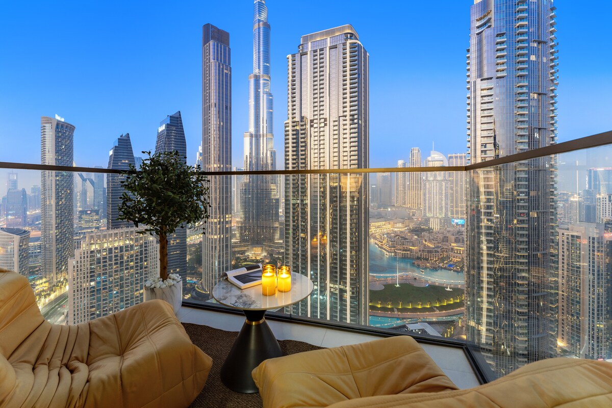 A balcony offers a stunning view of Downtown Dubai's skyline, featuring iconic buildings and the Burj Khalifa. Two modern lounge chairs are positioned around a small table, which holds lit candles. The ambiance is set against a twilight backdrop, highlighting the city's vibrant lights.