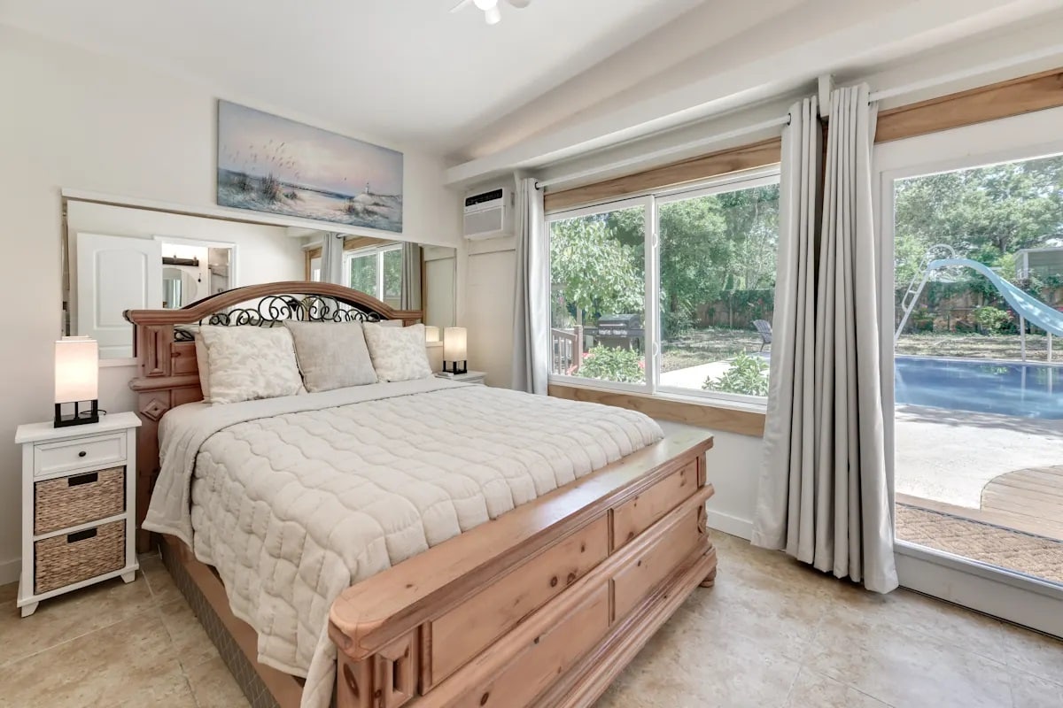 The spacious bedroom features a wooden bed frame with a quilted coverlet and two pillows. Natural light floods the room through large windows, revealing views of the outside area, including a pool and greenery. Accent lamps are placed on each side of the bed.