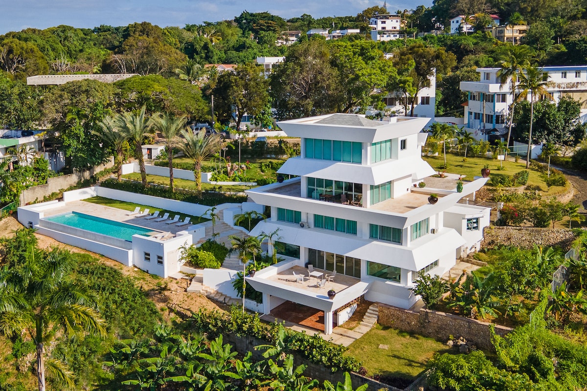 A modern four-story villa is showcased from an aerial perspective, featuring expansive terraces and a private pool surrounded by lush landscaping. The structure is characterized by a sleek design, large windows, and a spacious layout, all set against a backdrop of greenery and the distant hills.