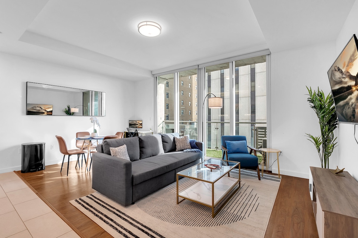 A spacious living area showcases a gray sofa arranged with various cushions, complemented by a glass coffee table. Natural light floods through large windows, illuminating a dining table for four and a comfortable blue chair. A flat-screen TV is mounted on the wall, enhancing the modern ambiance.