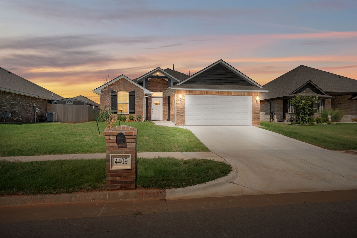 A modern home is shown from the street, featuring a well-maintained lawn and a brick exterior. The entrance is highlighted by archways and large windows, while the three-car garage provides ample parking space. Soft evening light enhances the welcoming atmosphere of the property.