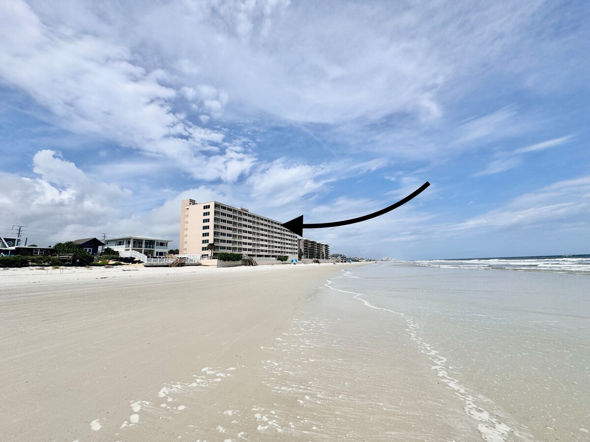 An expansive view of a wide, traffic-free beach features soft, light sand meeting gentle ocean waves. In the background, a multi-story building stands prominently, offering direct oceanfront access. The sky above displays fluffy clouds under a bright blue expanse.