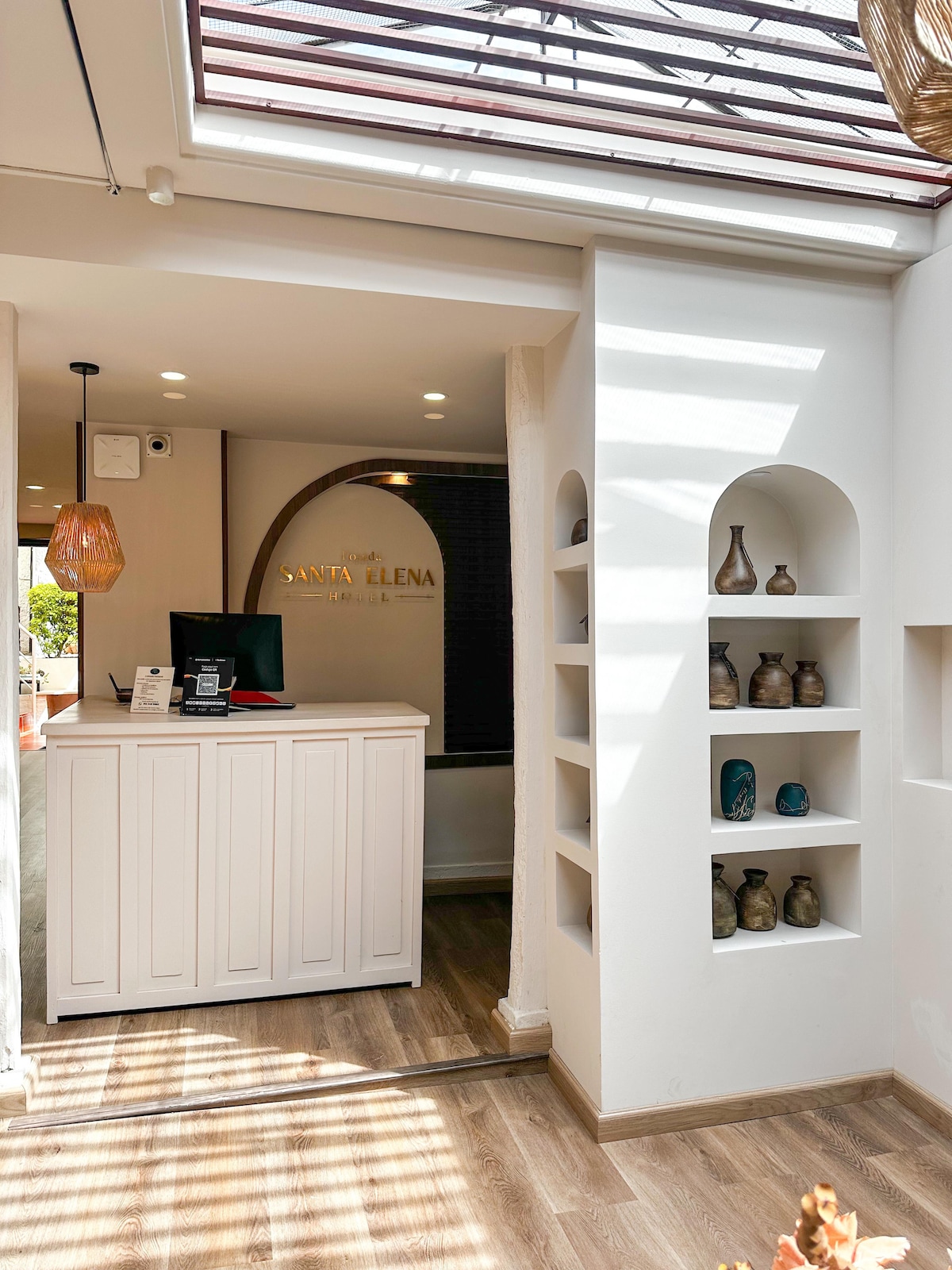 The reception area is characterized by a modern design with a white reception desk and a welcoming ambiance. Natural light enters through a skylight, illuminating decorative shelves showcasing a variety of vases and unique art pieces, enhancing the overall aesthetic of the space.