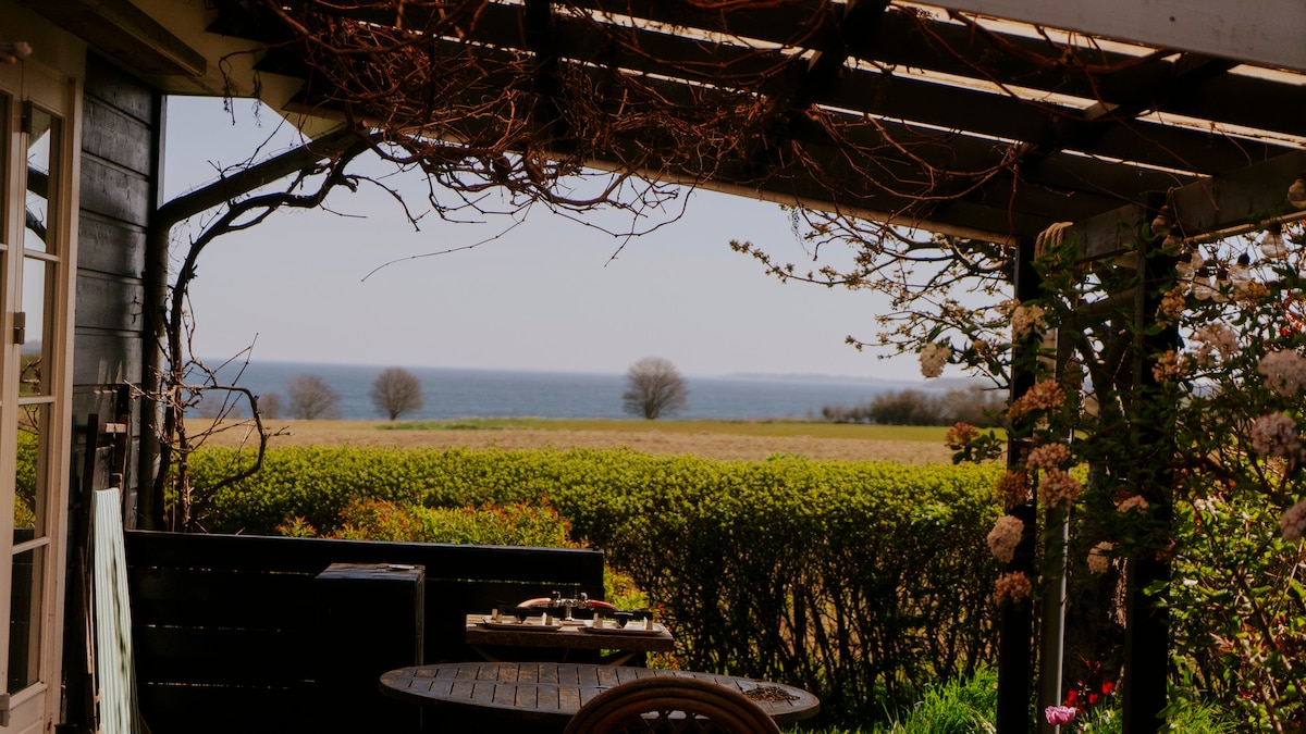 A shaded patio area is visible, featuring a round table and chairs surrounded by greenery. The view beyond includes a serene landscape leading to a body of water, framed by trees and a clear sky.