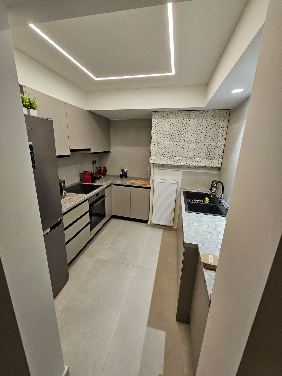 A modern kitchen is visible, featuring sleek cabinetry and a countertop with a subtle stone pattern. Stainless steel appliances are positioned, including a refrigerator and an oven. Natural light is softly diffused through a patterned window covering, enhancing the clean and organized space.