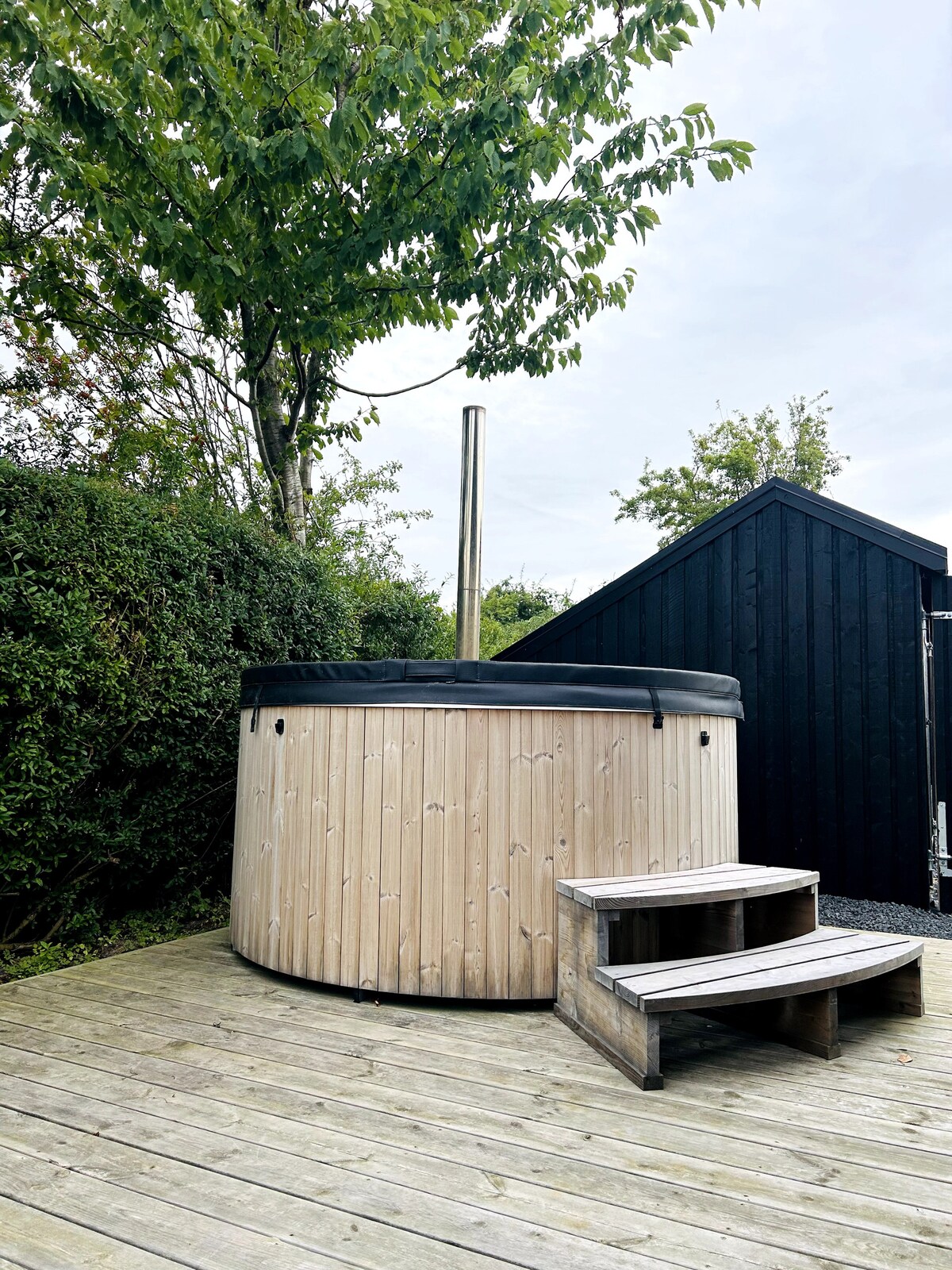 A wooden hot tub is featured on a wooden deck, surrounded by lush greenery. Steps lead up to the tub, while a black structure is visible in the background. The atmosphere is one of relaxation and natural tranquility.