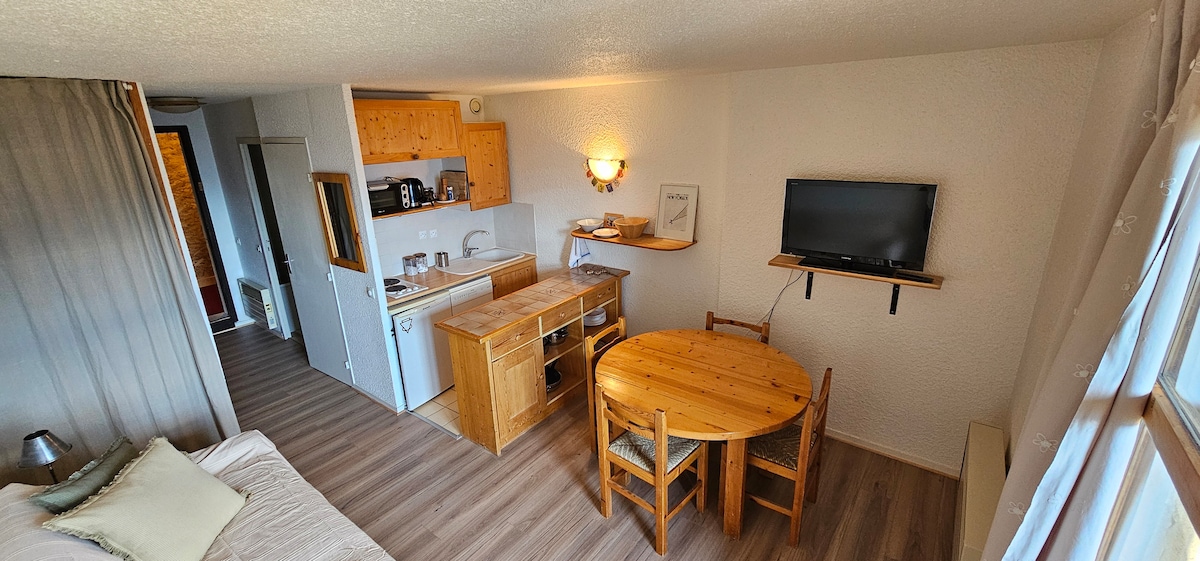 A bright living space includes a wooden dining table with four chairs, and a compact kitchen area equipped with modern appliances. Natural light enters through large windows, illuminating the warm wood tones and light walls, creating a cozy environment.