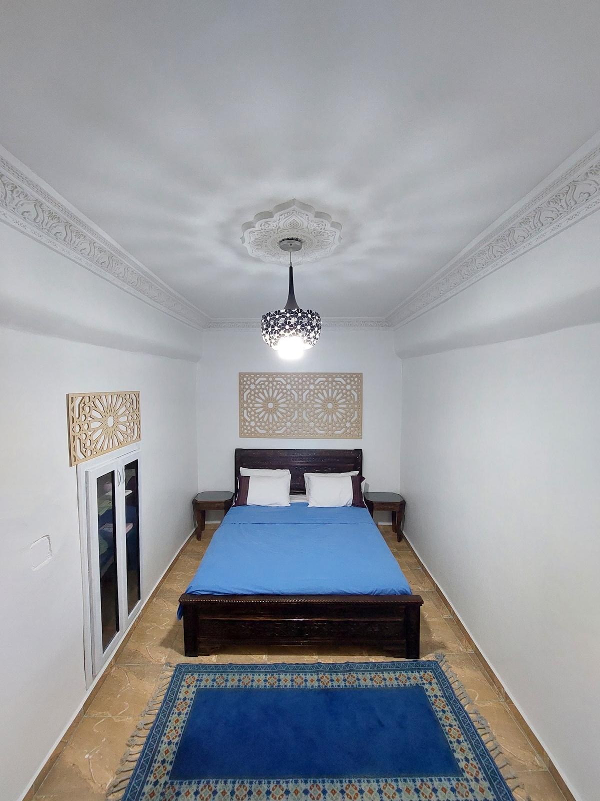 A bright bedroom is furnished with a wooden bed dressed in a blue coverlet. Two white pillows rest on the bed, and a decorative rug is placed on the floor. A light fixture hangs from the ceiling, casting a soft glow in the space.
