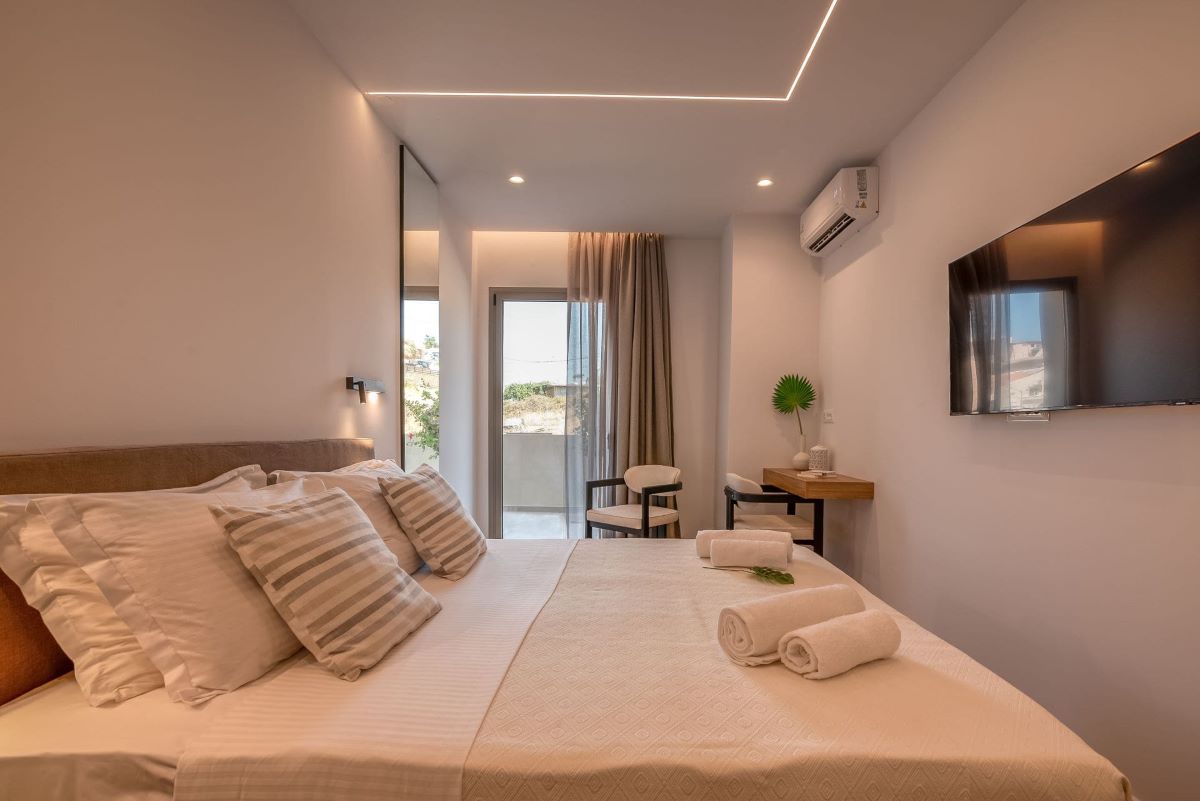 A cozy studio is presented with a spacious bed featuring crisp white linens and decorative cushions. A sleek flat-screen TV is mounted on the wall, while natural light enters through glass doors leading to a private terrace. Minimalist furnishings, including a desk and chair, complete the space.