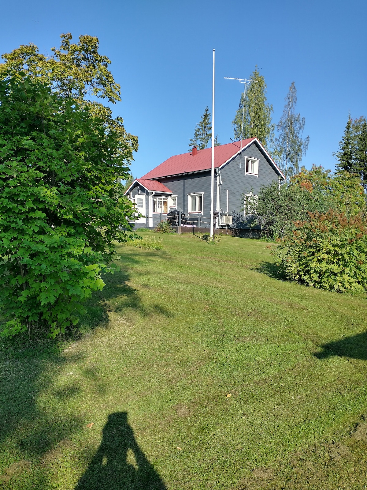 A two-story cabin with a red roof is situated on a spacious green lawn. Surrounding trees and shrubs create a natural ambiance, while the clear blue sky provides a bright backdrop. A flagpole stands in front, enhancing the outdoor setting.