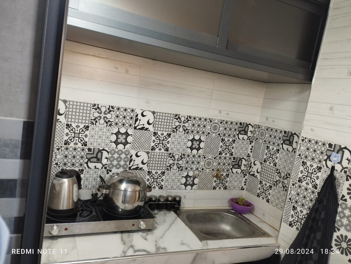 The small kitchen features a sleek design with patterned black and white tiles. A stainless steel kettle and pot rest on the stove, while a sink is positioned nearby, accompanied by a decorative bowl of fruit.