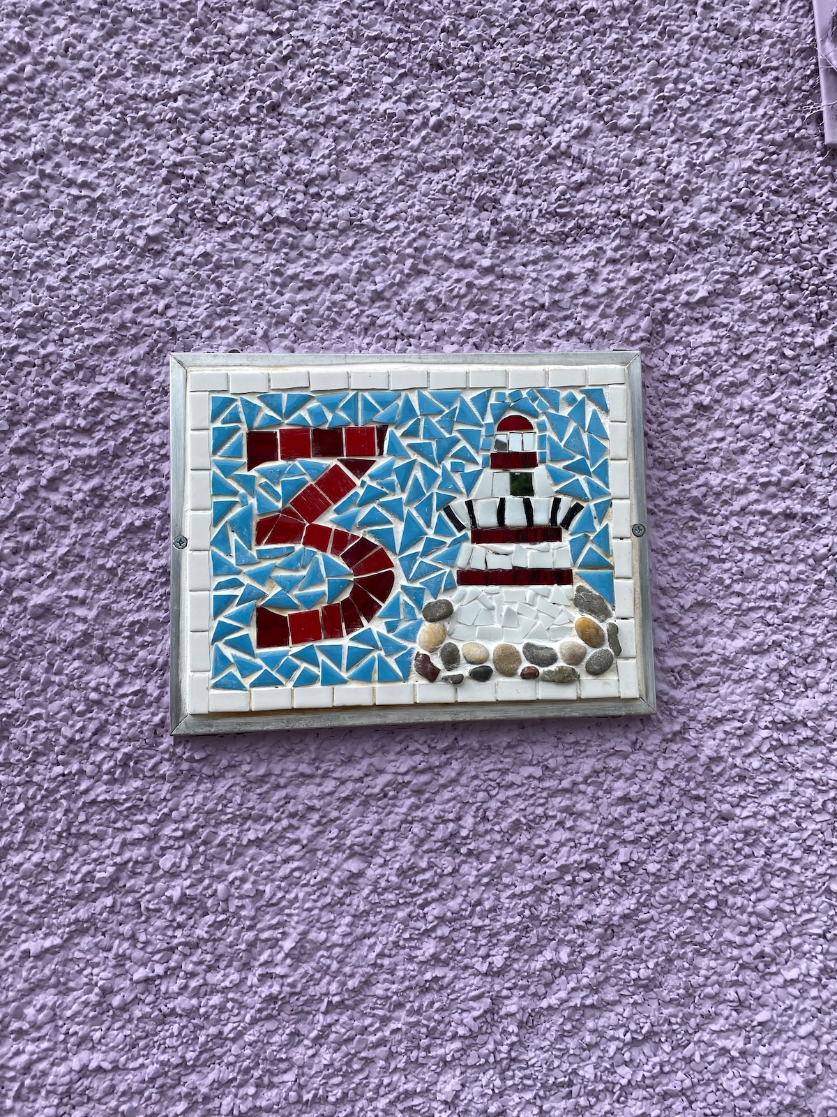 A decorative house number is displayed on a lavender wall. The tile design features the number '3' prominently, alongside a lighthouse illustration made from colorful mosaic pieces and small stones.