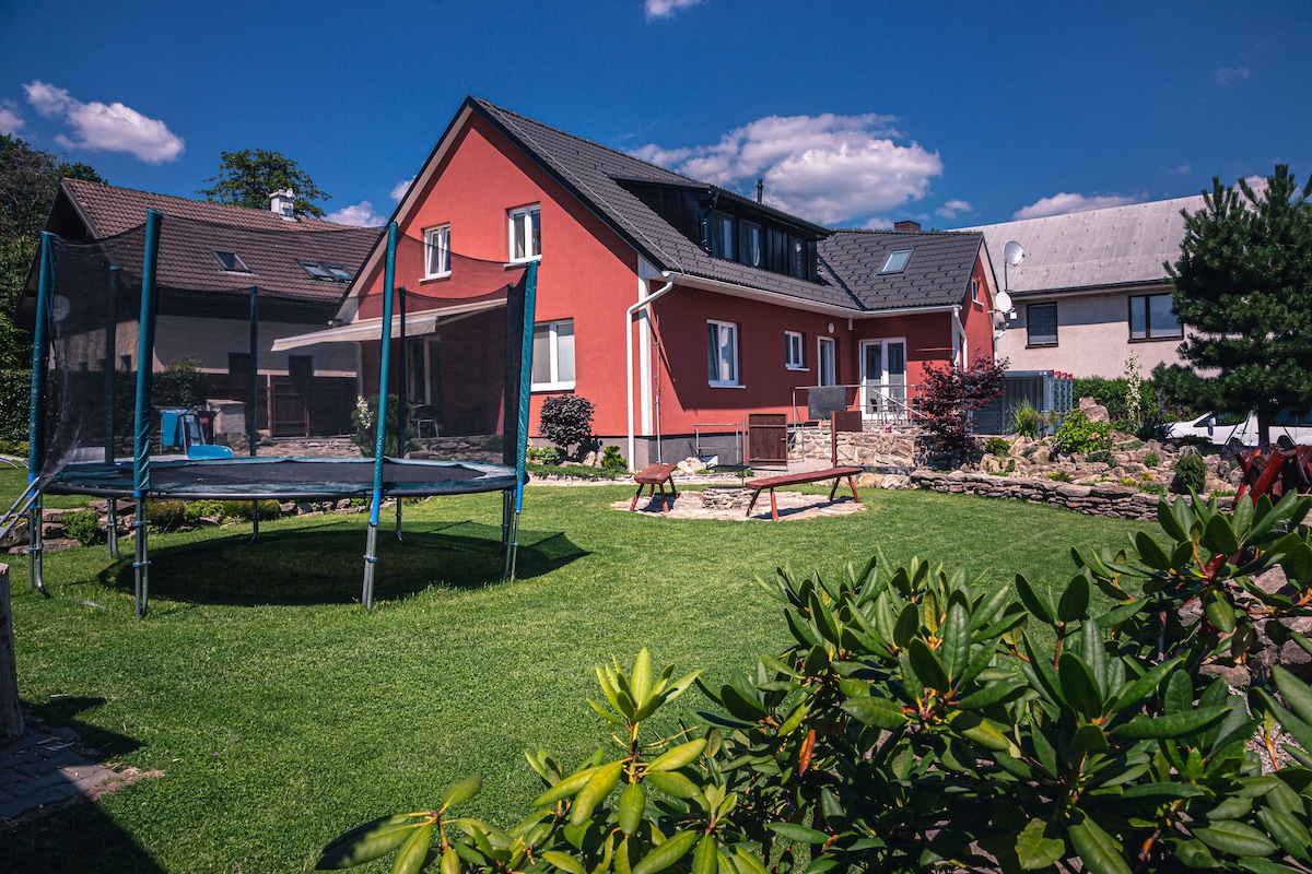 A spacious garden area surrounds a modern building, featuring a trampoline and picnic tables. Lush greenery and decorative plants enhance the outdoor space, with a clear blue sky and clouds providing a serene backdrop.