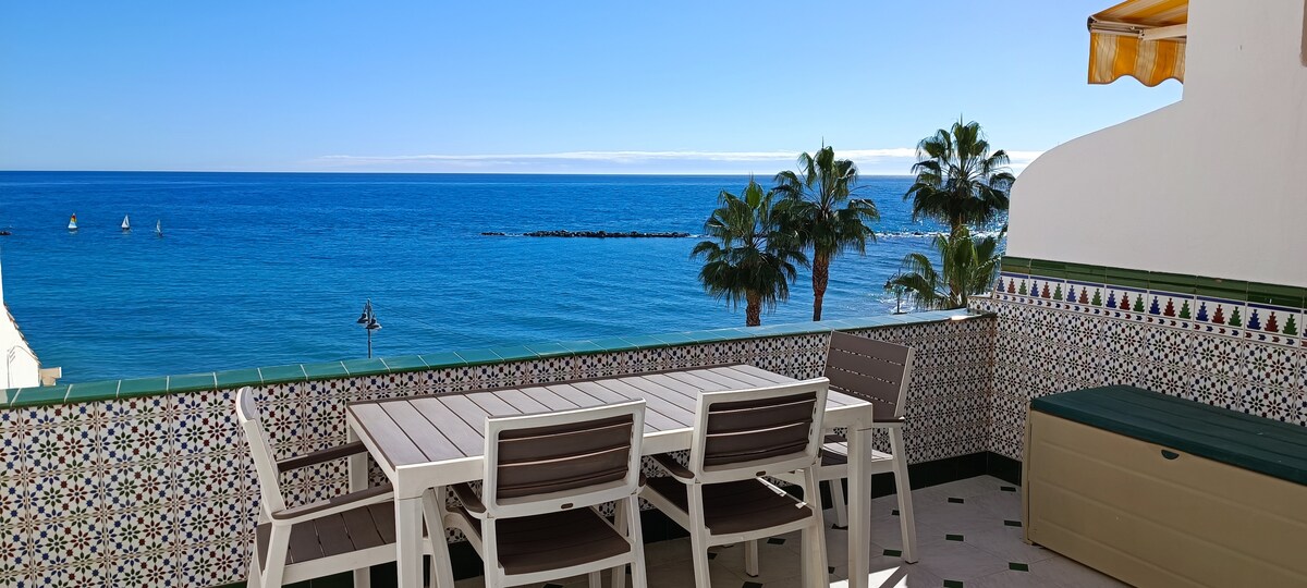 A terrace offers stunning views of the sea, with a dining table and several chairs arranged for outdoor seating. The backdrop includes palm trees gently swaying and the expansive blue ocean, creating a serene coastal ambiance.