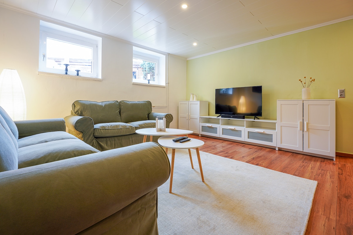 The spacious living area is furnished with a comfortable green sofa set, complemented by a modern coffee table. A large flat-screen TV is positioned on a white media console. Natural light enters through two small windows, illuminating the hardwood floor and soft area rug.