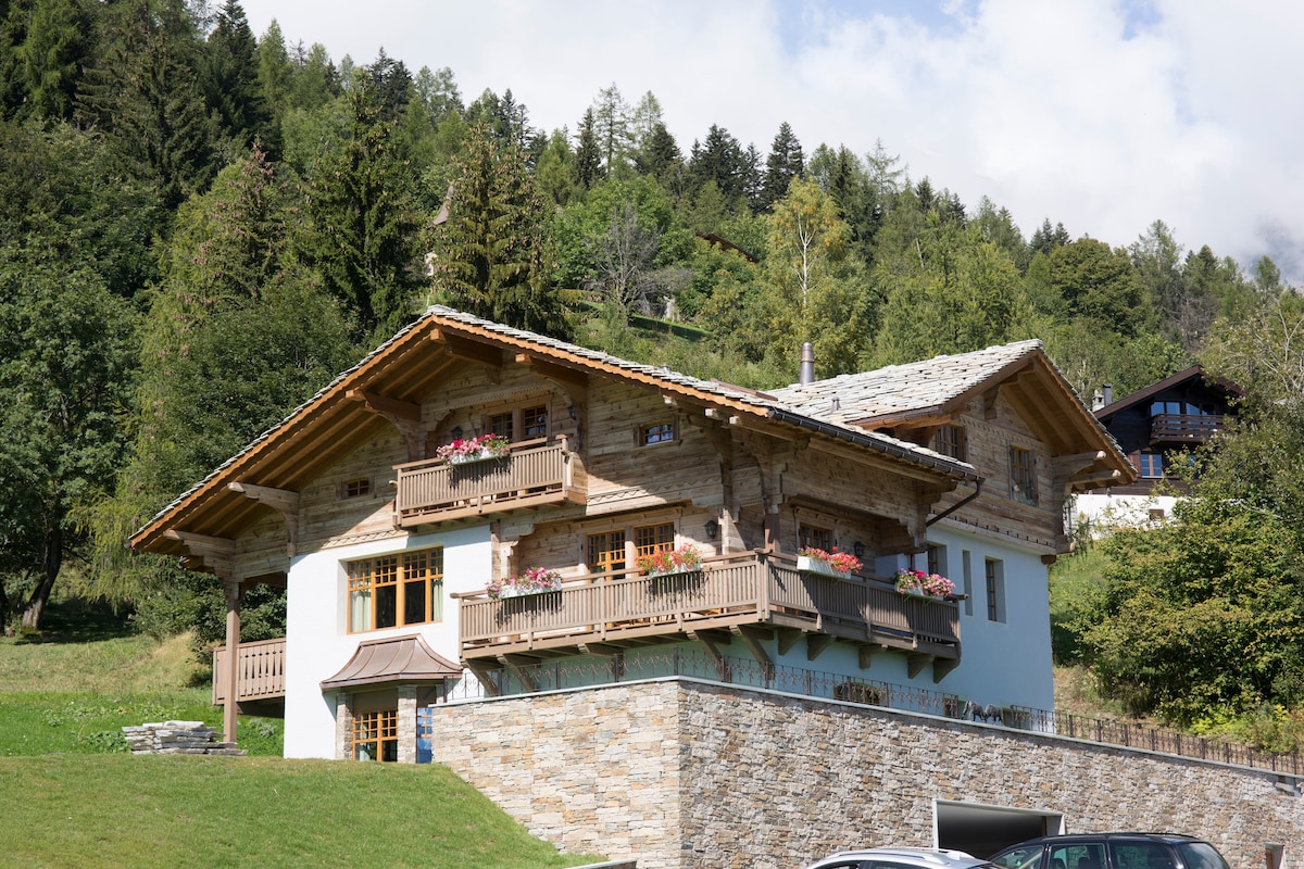 The chalet is situated amidst lush greenery, showcasing a wooden façade accentuated by balconies adorned with flower boxes. A stone wall wraps around the ground level, complementing the rustic charm. Tall trees are visible in the background, enhancing the serene mountain setting.