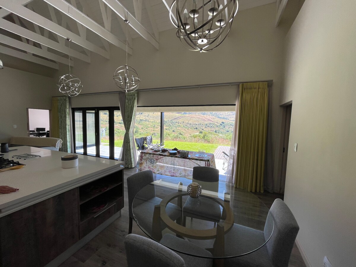 An open layout kitchen area is highlighted by a round glass table surrounded by four upholstered chairs. Large glass doors provide a view of the outdoor space, where natural scenery can be seen in the background, brightened by ample natural light.
