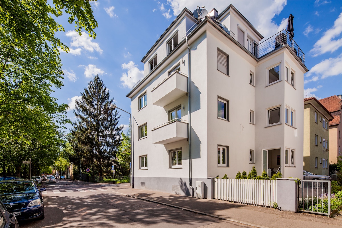 A modern three-story building is situated on a tree-lined street. The exterior is characterized by clean white walls and multiple balconies. Surrounding greenery complements the structure, and a white picket fence adds a welcoming touch to the property.