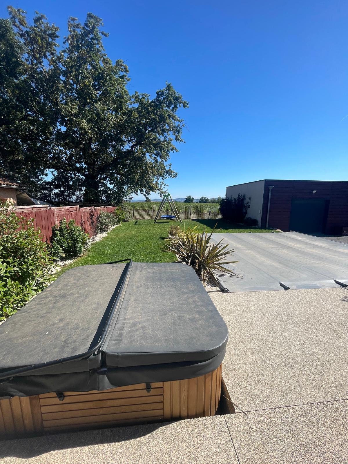 A spacious outdoor area is shown, featuring a hot tub with a protective cover. Lush green grass extends beyond the tub, accompanied by a swing set in the distance. The landscape is bordered by trees under a clear blue sky.