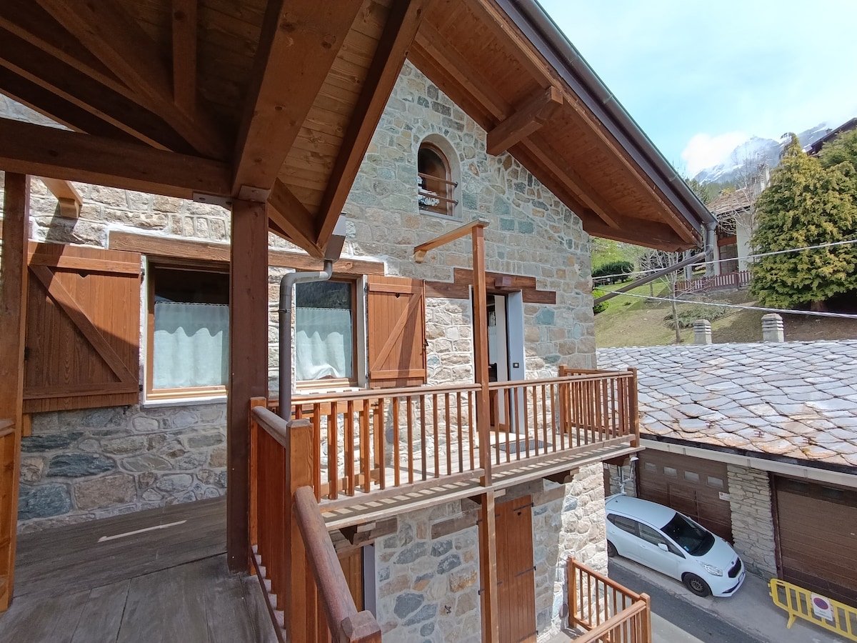 The exterior features a charming stone facade with wooden accents and window shutters. A private balcony offers outdoor space, while the sloped roof and stonework contribute to a rustic aesthetic. Surrounding greenery and a glimpse of the mountain landscape are visible in the background.