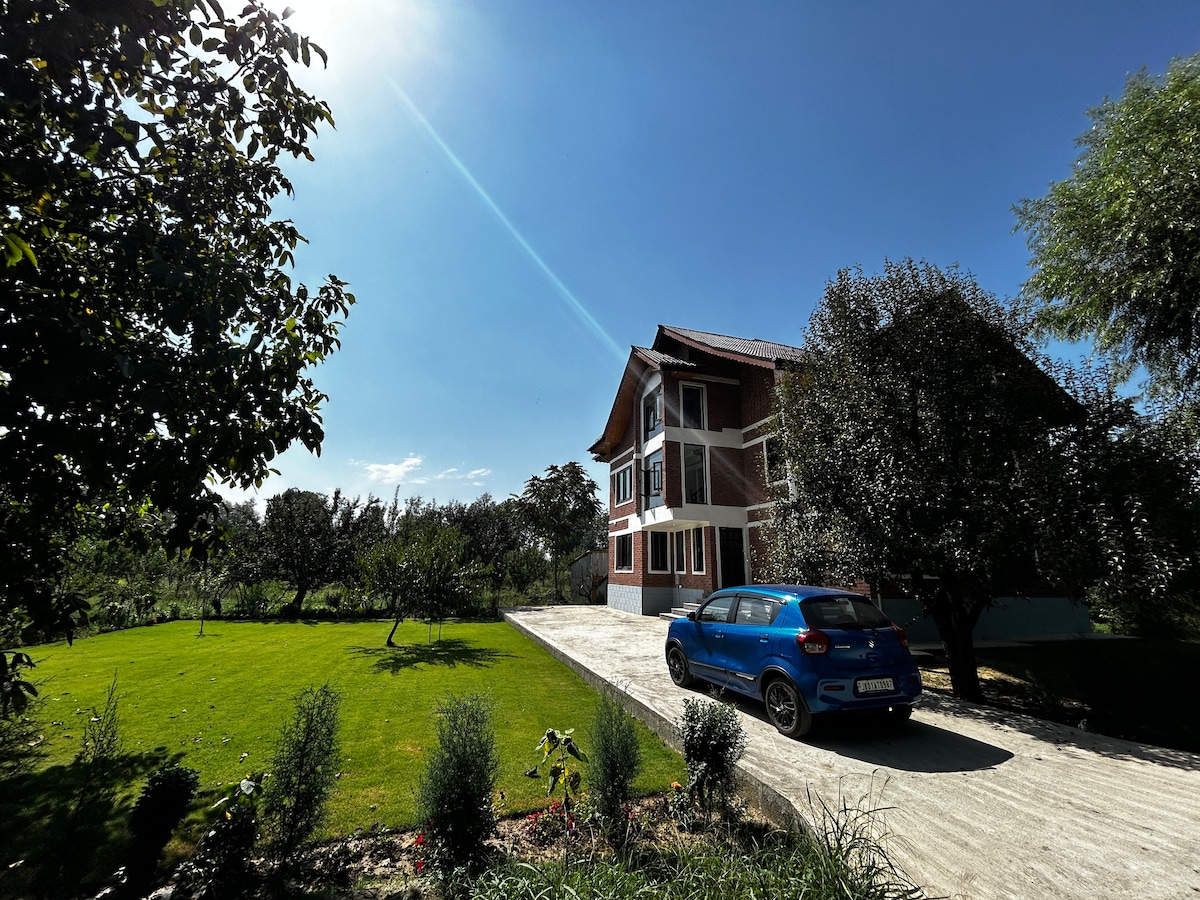 The exterior of the cottage is framed by a vibrant green lawn and fruit trees. A well-maintained driveway leads to the entrance, with a blue car parked nearby. The building features multiple stories and large windows, reflecting the clear blue sky.