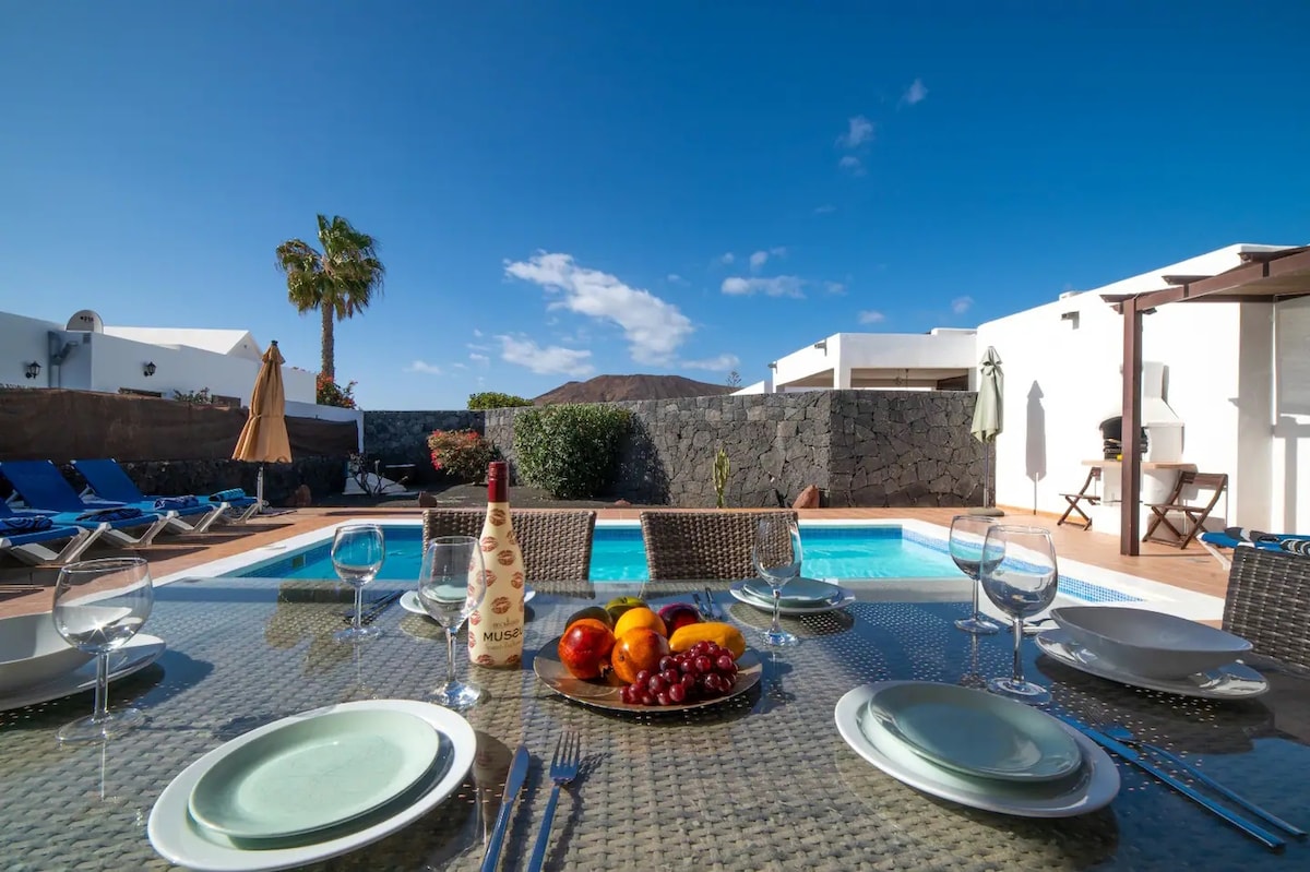 An outdoor dining area is showcased with a large, glass-topped table set for a meal. A bowl of fresh fruits sits at the center, complementing the inviting private heated pool and sun loungers nearby. Lush greenery and a clear blue sky frame the scene.