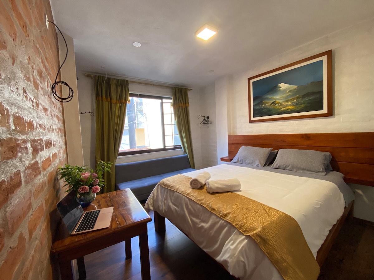 A cozy room is equipped with a double bed featuring soft linens and a decorative golden throw. A window provides natural light and a view of the outside. A small table holds a laptop and a vase with fresh flowers, while a seating area is present beside the window.