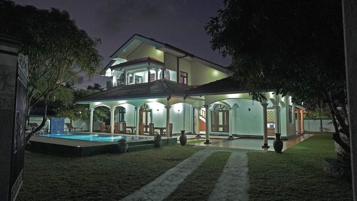 The villa's exterior is highlighted in soft lighting, featuring a welcoming façade with arched doorways and a spacious terrace. A tranquil pool reflects the surrounding greenery, while a pathway leads through the well-maintained lawn, offering a glimpse of the inviting atmosphere.