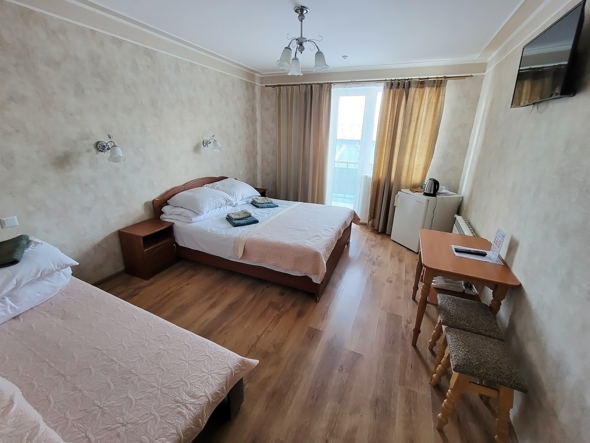 A bright and airy room features two spacious beds, each dressed in light linens. Natural light enters through a large window dressed with sheer curtains. A small table and two stools are positioned nearby, alongside a kettle and refrigerator for guest convenience.