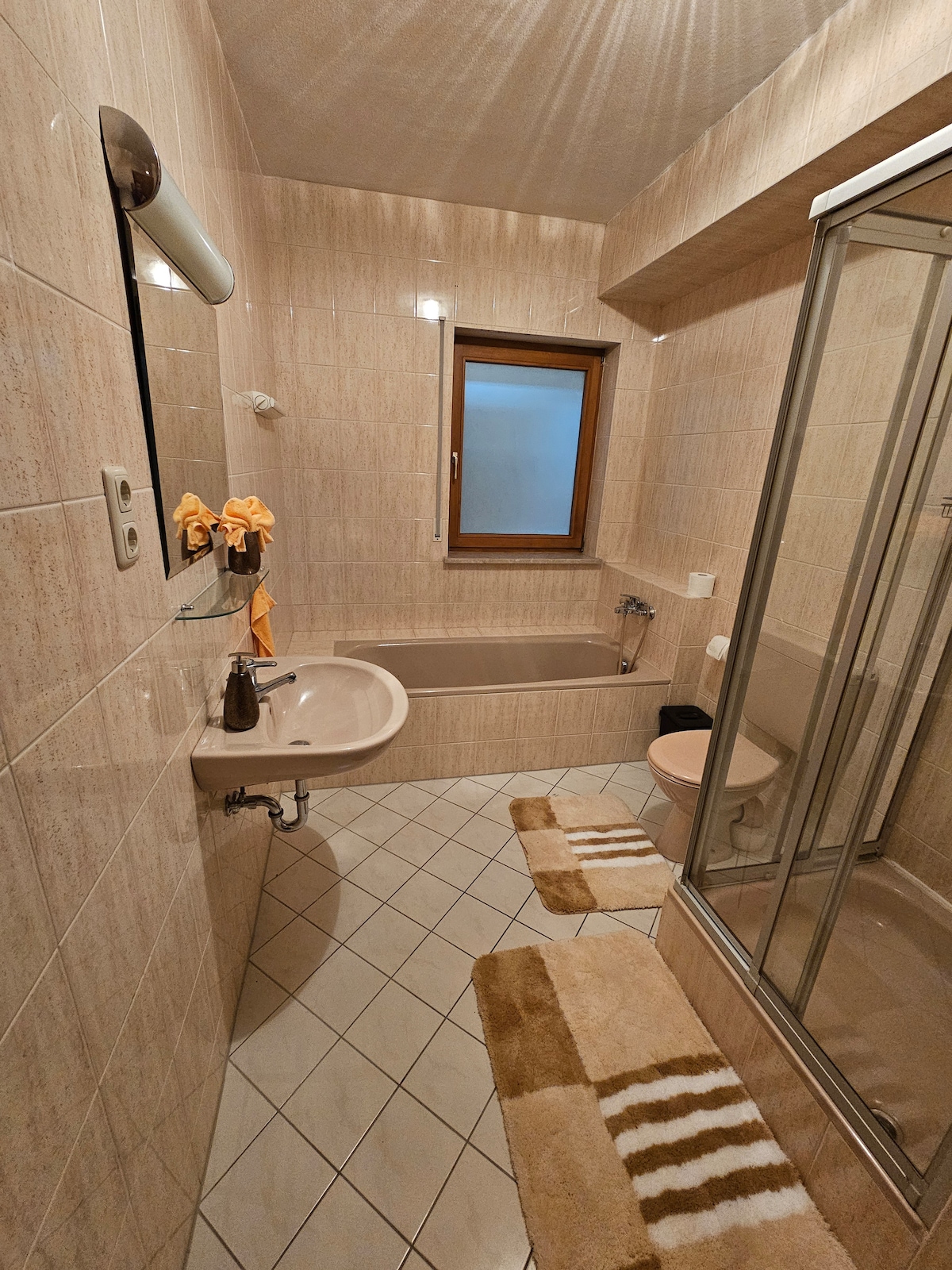 The bathroom features beige tiled walls and a window allowing natural light. It includes a shower stall, a bathtub, a sink with a mirror above, and a toilet. Soft, striped bath mats are placed on the tiled floor for comfort.