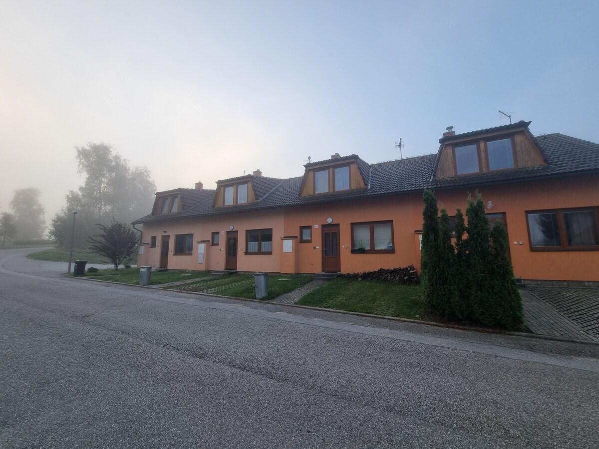 A row of modern buildings is seen along a quiet street, with a layer of mist creating a serene atmosphere. Each building features large windows and a mix of orange and brown facades, with greenery enhancing the setting.