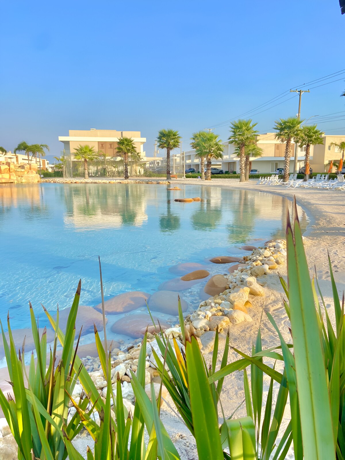 A serene artificial beach features calm, clear water surrounded by soft sandy edges and decorative stones. Lush green palm trees line the water's edge, casting reflections on the tranquil surface, while lounge chairs are arranged nearby for relaxation.
