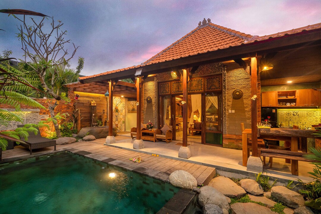 A luxury private villa is set in a tropical environment, featuring a serene pool surrounded by natural stones and lush greenery. The open design creates a seamless connection between living spaces, kitchen, and outdoor area. The warm glow of evening lights adds an inviting charm.