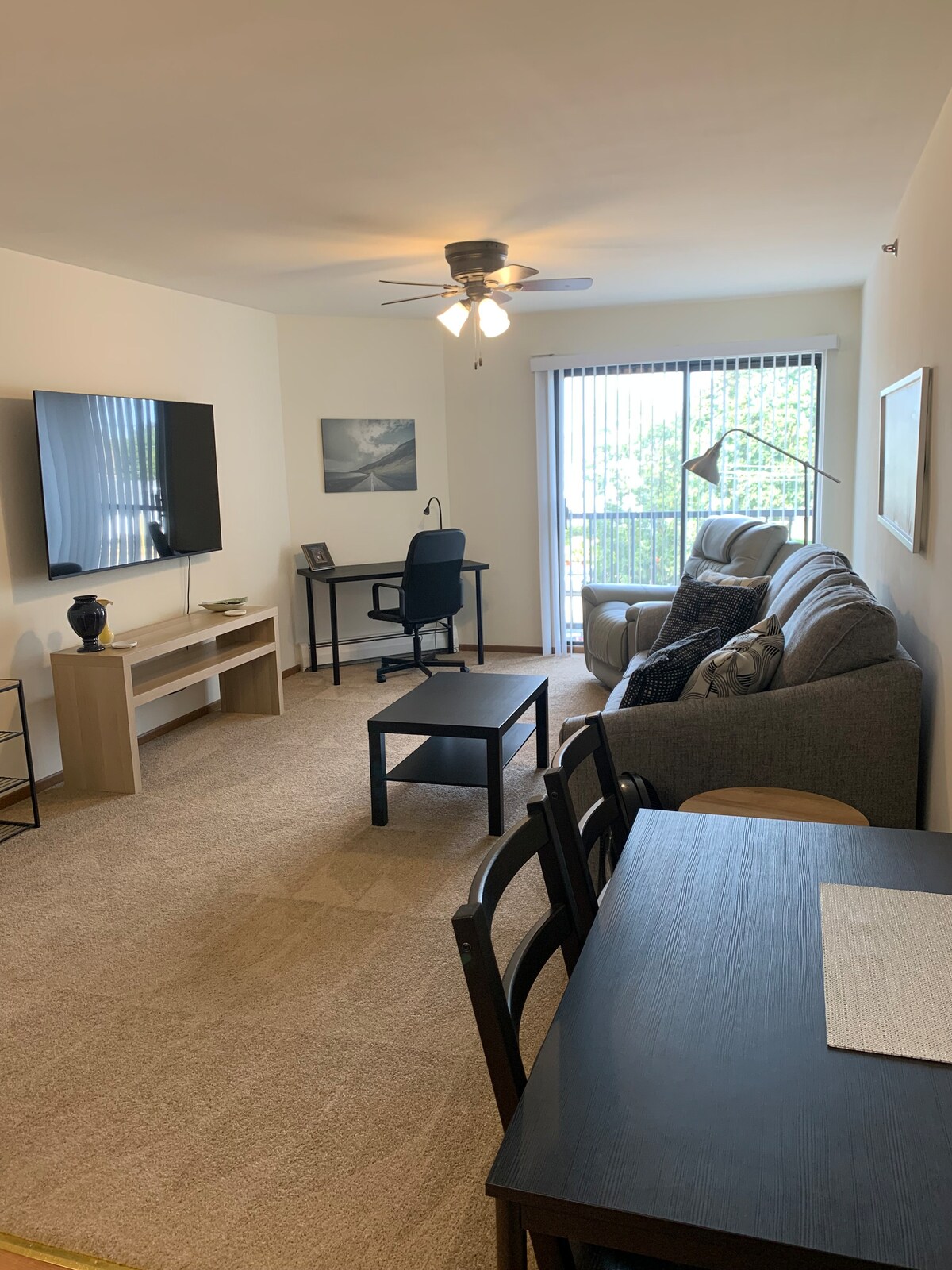 A comfortable living area is designed with a gray sofa and a black coffee table, complemented by a dining table. A work desk is positioned near large windows, allowing natural light. A ceiling fan is visible, and a flat-screen TV is mounted on the wall.