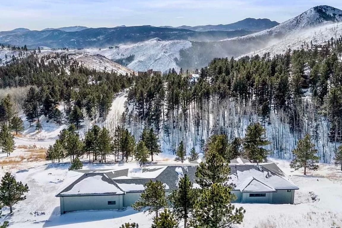 A scenic view of a mountain retreat surrounded by snow-covered trees and hills. The landscape showcases a vast expanse of nature, with peaks in the background and a serene, winter atmosphere. The structure is partially visible, harmonizing with the natural surroundings.