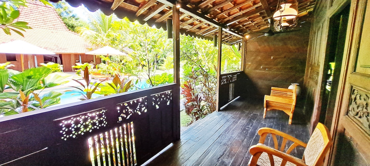 An inviting porch is presented with dark wooden flooring and decorative wooden railings. Two wooden chairs offer seating, surrounded by lush tropical foliage. In the background, glimpses of the swimming pool and nearby villas can be seen, creating a serene outdoor space.