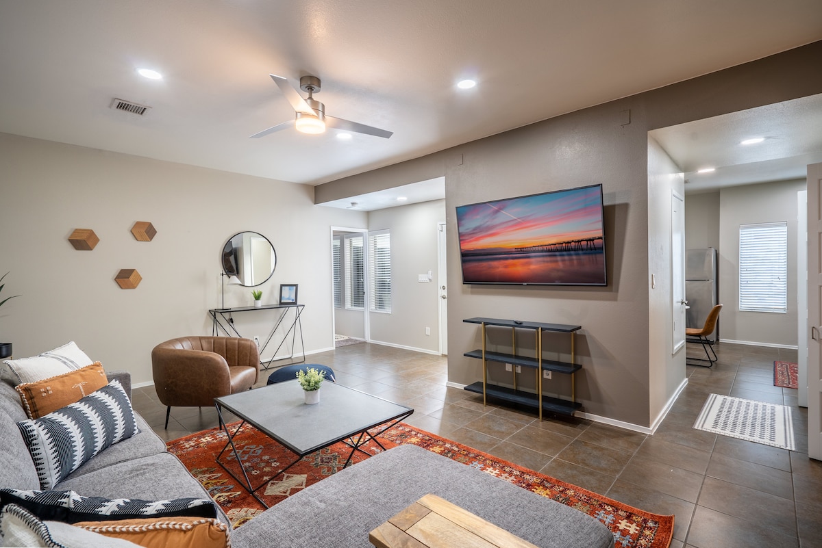 A spacious living area features a comfortable sectional sofa, a wooden coffee table, and a flat-screen television mounted on the wall. Natural light filters through the nearby windows, highlighting decorative elements on the walls and the warm tones of the tiled floor.