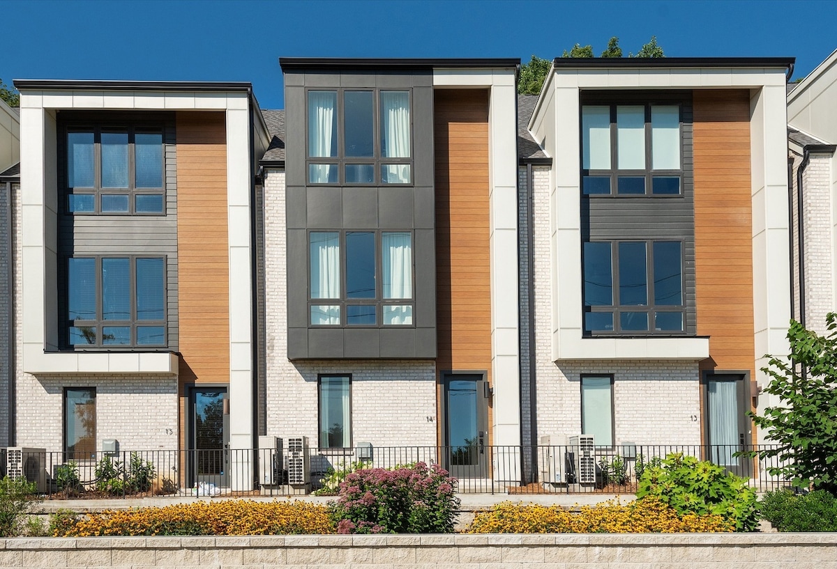 A modern multi-story building is showcased with a blend of wood and brick exteriors. Large windows reveal light curtains that complement the clean lines of the architecture. Flowering shrubs and greenery adorn the front, enhancing the inviting entrance.