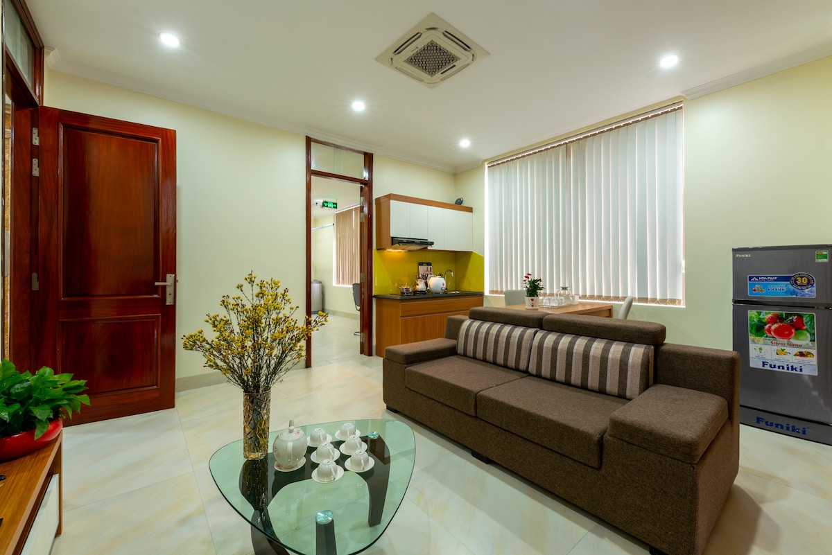 A modern living area is highlighted by a brown sofa with striped cushions, positioned across from a glass coffee table. Natural light enters through large windows with white blinds, illuminating the space. A kitchenette is visible in the background, featuring minimalistic cabinetry.