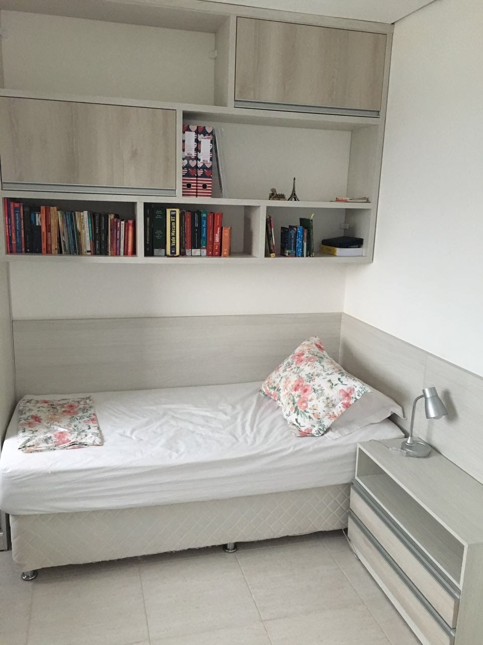 A compact sleeping area features a neatly made single bed with floral accent pillows. Above, shelves are lined with books and decorative items. A small bedside table with a lamp is positioned next to the bed, contributing to the room's organized appearance.