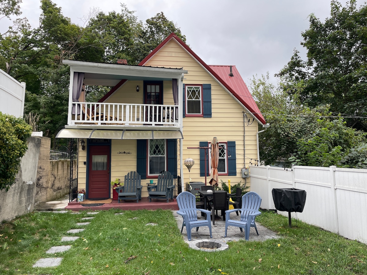 A charming two-story house features yellow siding and a red metal roof. The welcoming front porch is furnished with chairs, while a cozy outdoor seating area with a fire pit and grill is visible in the yard. Lush greenery surrounds the property.