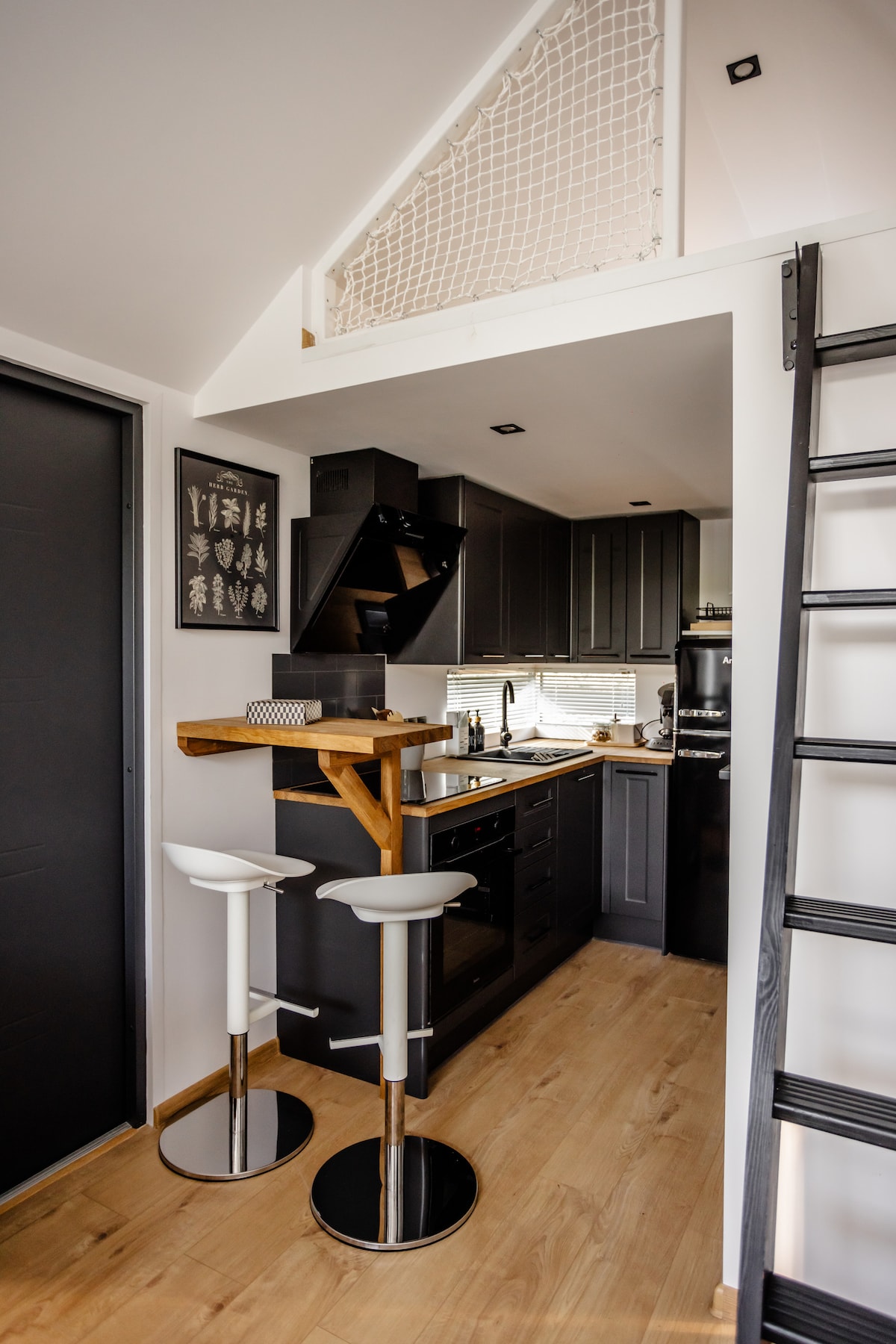 A modern kitchen area features sleek black cabinetry and stainless steel appliances. Bar stools are positioned at a small wooden counter, enhancing the space's functionality. Natural light filters through nearby windows, creating an inviting atmosphere.