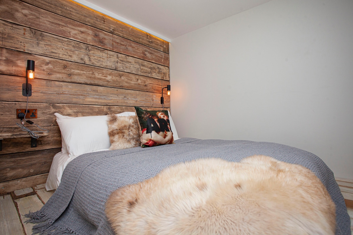 A cozy sleeping area features a spacious bed against a rustic wooden accent wall. The bedding is complemented by a textured gray throw and a soft fur blanket, with warm lighting provided by stylish wall sconces.