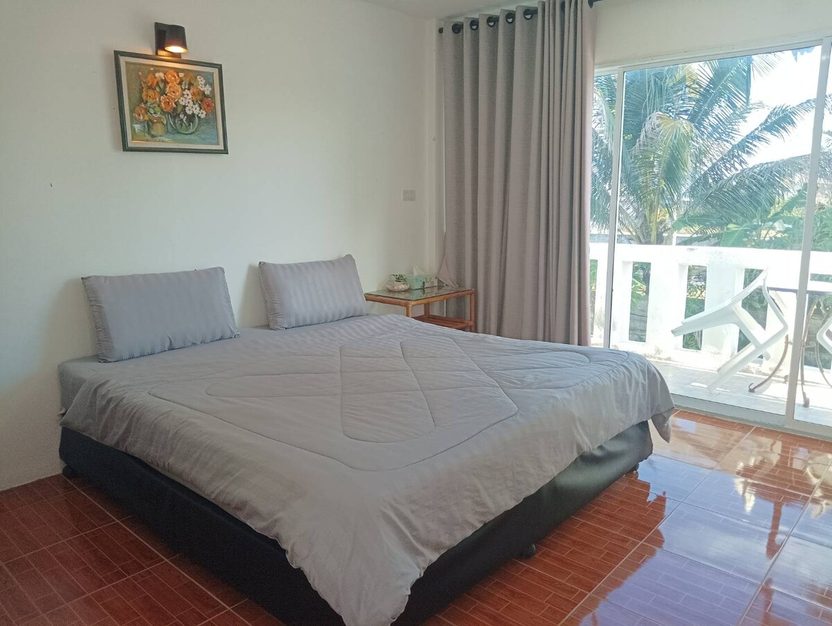 A spacious bed is centered in the room, dressed in a solid grey cover. Large windows allow natural light to fill the space, revealing a view of palm trees outside. A small table is positioned nearby, complemented by a decorative floral artwork on the wall.