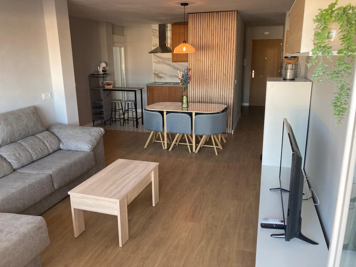 A modern living space is presented, featuring a comfortable gray sofa and a light wooden coffee table. A dining table with chairs is visible in the background, adjacent to an equipped kitchen with a hood. Soft lighting enhances the open-plan layout.
