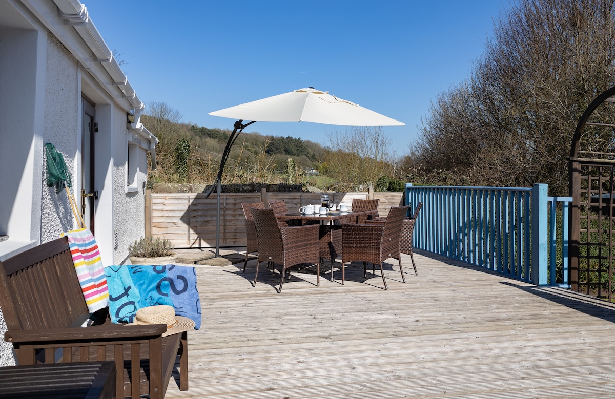 A large deck area features a table with seating for six, shaded by a parasol. A cozy chair with a colorful towel draped over it adds to the outdoor space, surrounded by a view of greenery and trees in the distance.