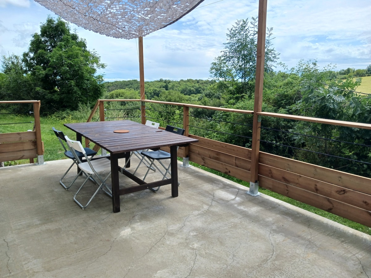 A spacious terrace features a wooden table accompanied by metal and fabric chairs, providing a comfortable outdoor dining area. A shaded overhead structure offers relief from sunlight, while greenery and distant hills are visible in the background, creating a serene setting.
