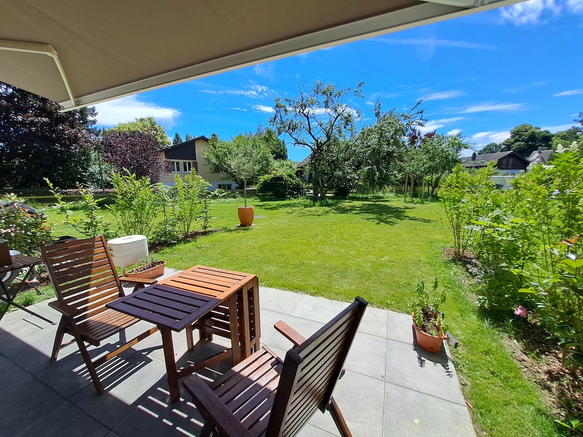 A serene outdoor space features a small wooden table and two chairs set on a stone patio. Lush greenery surrounds the area, with well-maintained grass and flowering plants. Bright blue skies enhance the peaceful atmosphere, making it an inviting space for relaxation.