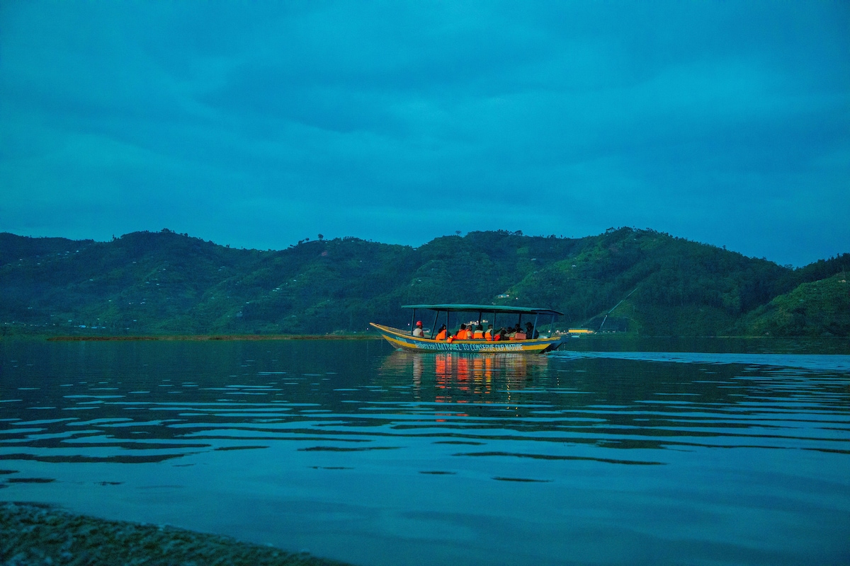 A boat is seen gliding across the calm waters of Twin Lakes, surrounded by lush green hills under a twilight sky. Gentle ripples reflect the boat's lights, enhancing the serene ambiance of the lakes.