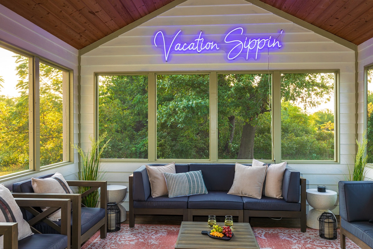 A bright three-season room is filled with natural light from large windows overlooking greenery. Comfortable seating is arranged with throw pillows on a dark sofa. A neon sign on the wall adds a playful touch, while a small table showcases refreshments.